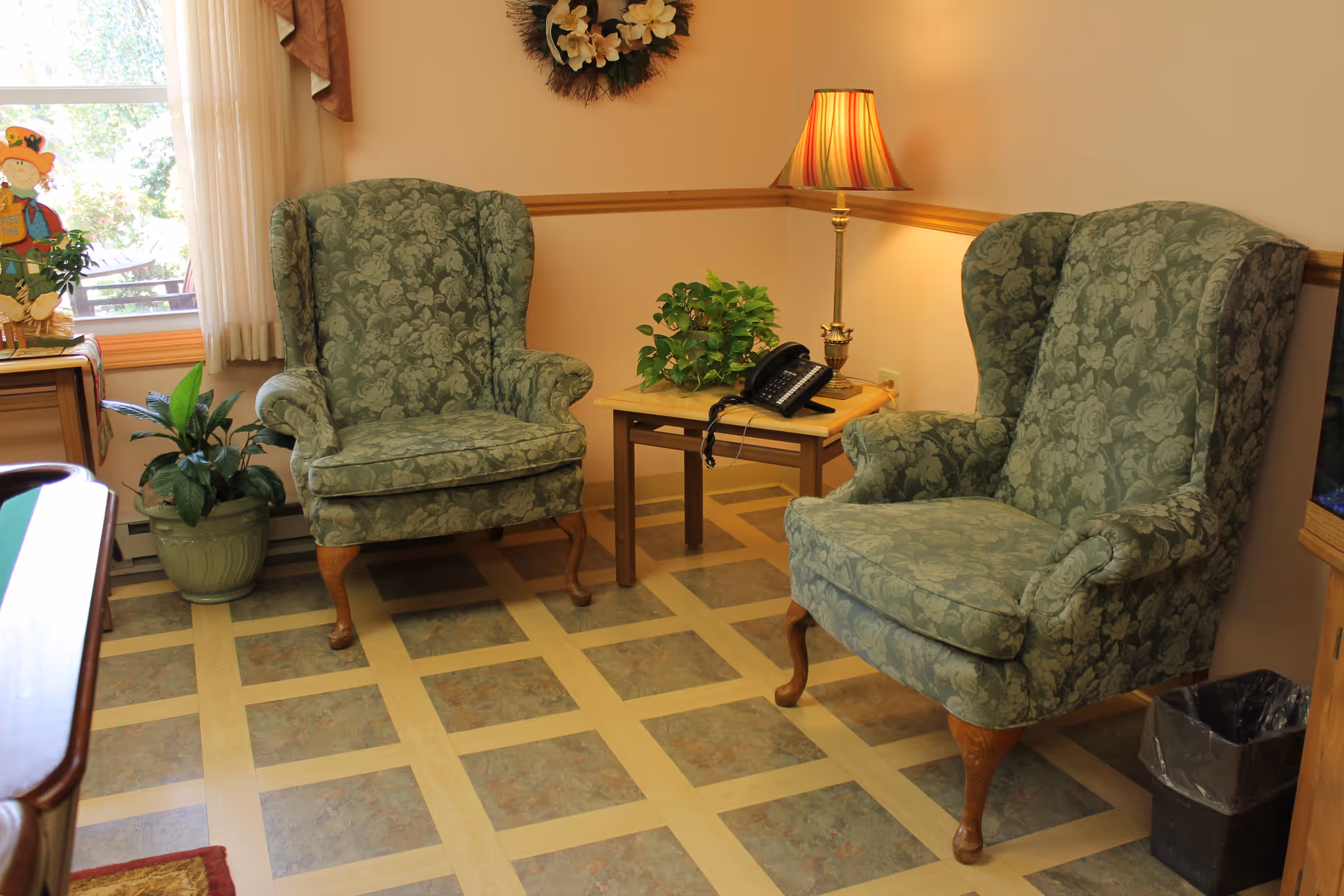 A cozy sitting area with two green floral upholstered armchairs facing each other. Between them is a small wooden table with a potted plant and a telephone. A lamp with a striped shade is on the table, casting warm light. There is a window with sheer curtains and a decorative wreath on the wall above the table. A potted plant is on the floor near the window, and a small trash bin is visible on the right side.
