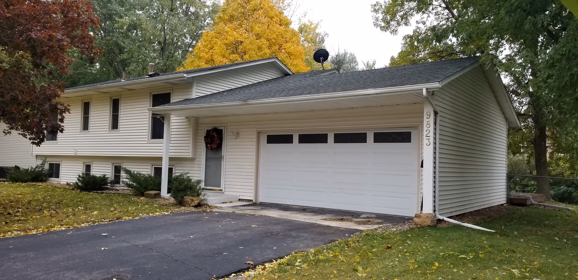 Front exterior of a light-colored split-level house with an attached two-car garage, driveway, and autumn trees.