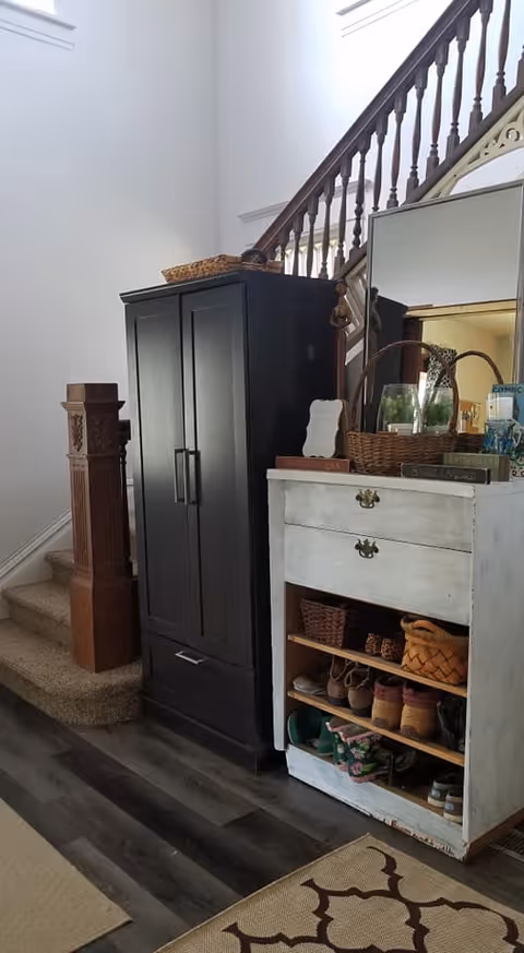 Interior view of a home entryway featuring a carpeted staircase with wooden banister, a tall black cabinet, and a white distressed dresser with shelves holding shoes and baskets. A large mirror is placed on top of the dresser along with decorative items including a wicker basket and books. The floor is dark wood with a patterned area rug partially visible.