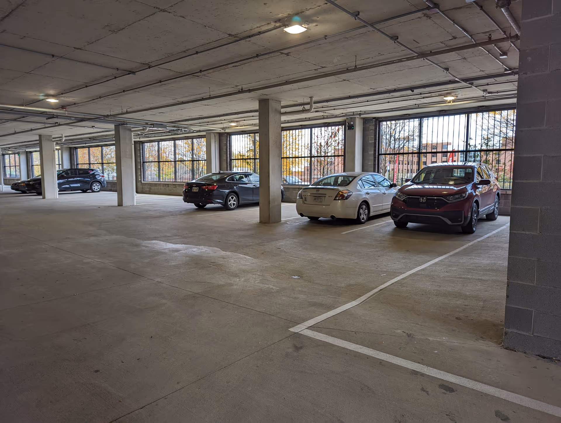 Indoor parking garage with several parked cars along the right side and large windows with metal bars letting in natural light.