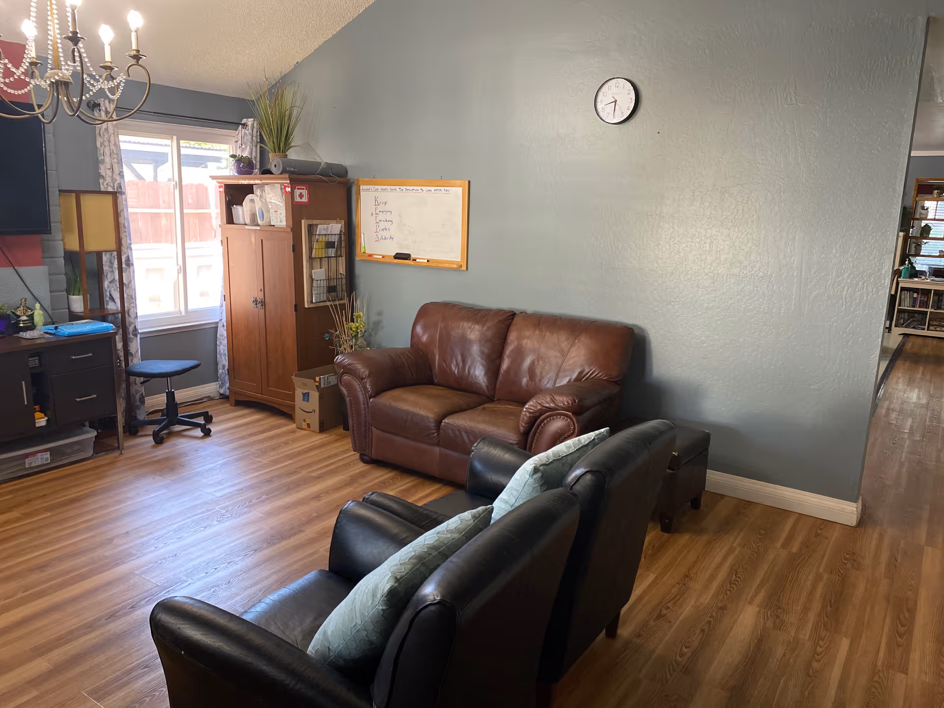 A cozy living room with a brown leather loveseat and two black armchairs with light blue pillows. There is a wooden cabinet near a window with curtains, a whiteboard on the wall, a clock above the loveseat, and a chandelier hanging from the ceiling. The floor is wooden and there is a hallway visible to the right.