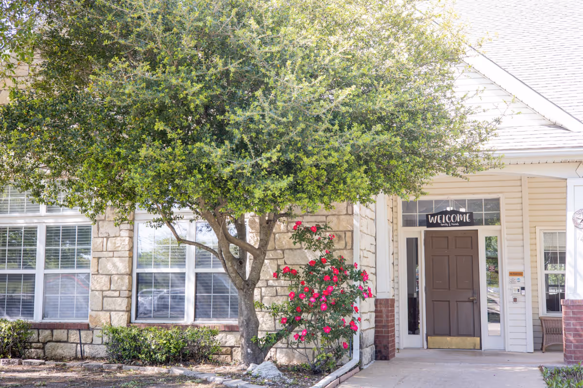 Entrance to a senior living building with a 'Welcome' sign above a dark brown door, stone exterior, a large tree, and pink flowering bushes.