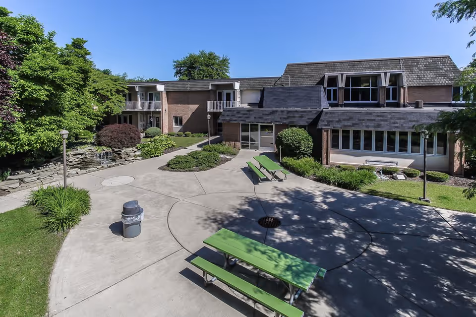 Sunlit courtyard with green picnic tables on a circular concrete patio in front of a two-story brick building.