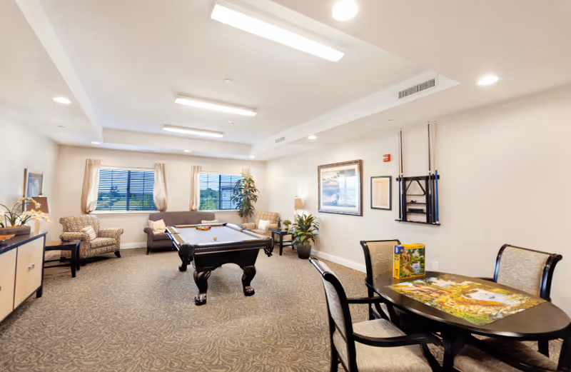 A well-lit recreational room with a pool table in the center, surrounded by comfortable armchairs and a sofa near windows with beige curtains. There is a round table with chairs and a puzzle on it in the foreground. The walls are decorated with framed pictures and a rack holding pool cues.