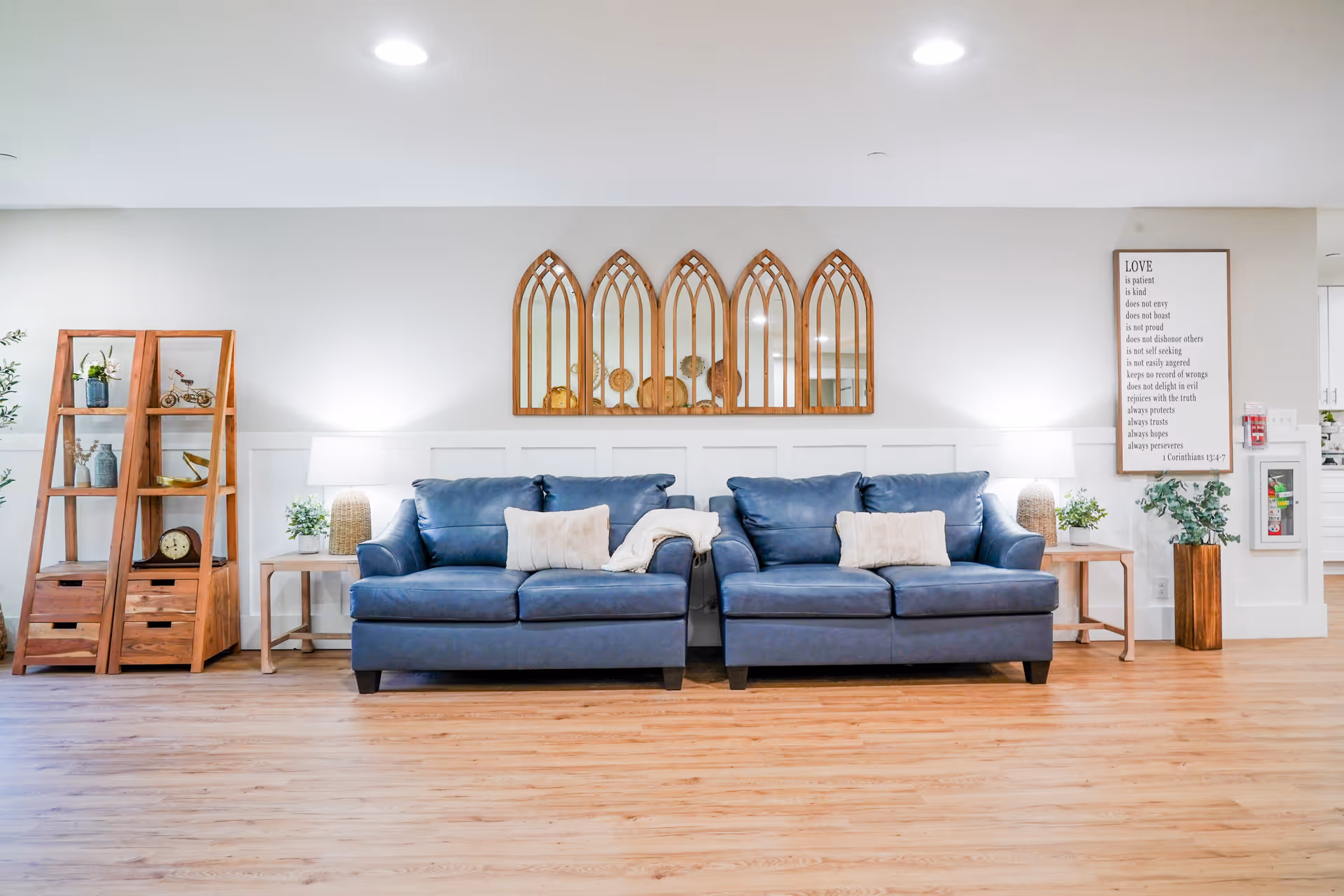 A bright and modern living room area featuring two blue leather sofas with cream-colored pillows and a throw blanket. Behind the sofas, there are five wooden framed mirrors with an arched design mounted on the wall. On either side of the sofas are wooden side tables with lamps and small plants. To the left, there are two wooden shelving units displaying decorative items including vases, a clock, and a small bicycle model. To the right, a framed inspirational quote is hung on the wall above a wooden planter with greenery. The room has light wood flooring and white walls with recessed ceiling lights.