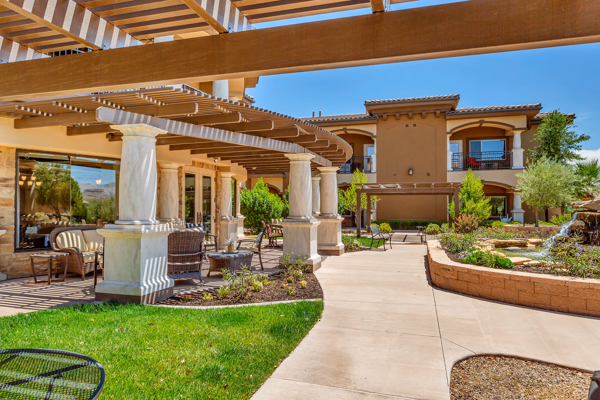 Outdoor patio area at The Retreat at Sunbrook featuring a covered seating area with wicker chairs and tables, marble columns supporting a wooden pergola, a paved walkway, green grass, landscaped garden beds, and a water feature with a small waterfall. The building has a tan exterior with balconies and large windows under a clear blue sky.