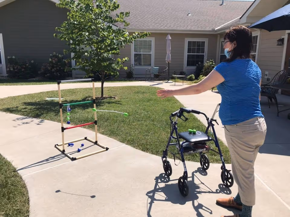 A woman wearing a blue shirt and beige pants is playing an outdoor ladder toss game on a paved area with grass and a small tree nearby. She is standing next to a walker and appears to be throwing bolas towards the ladder target. The setting is outside a residential building with windows and patio furniture visible.