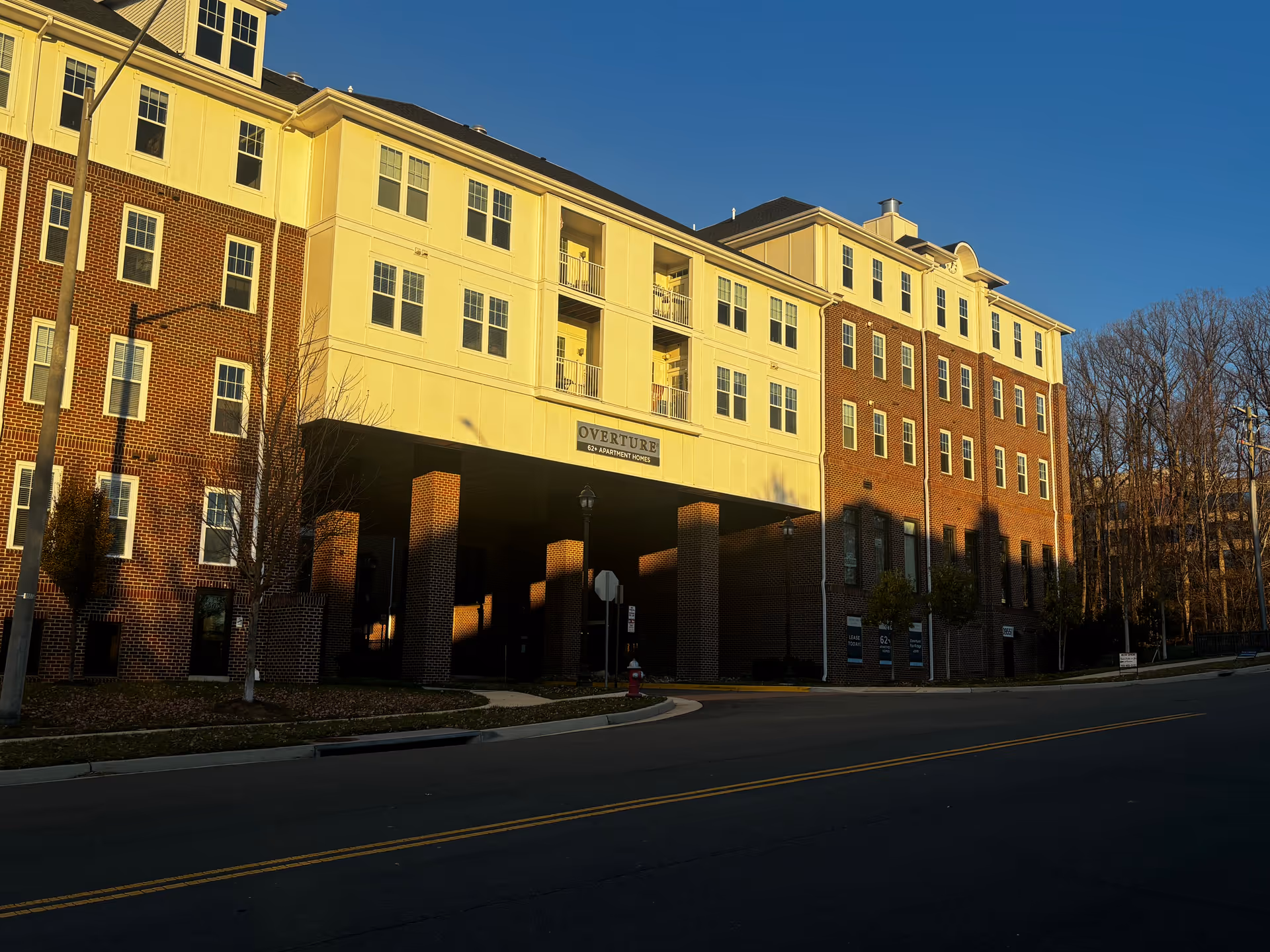 Exterior view of a multi-story senior living facility building named Overture Fair Ridge, with a combination of brick and light-colored siding, multiple windows, balconies, and a covered entrance area. The scene is captured during sunset with clear blue sky and some leafless trees in the background.