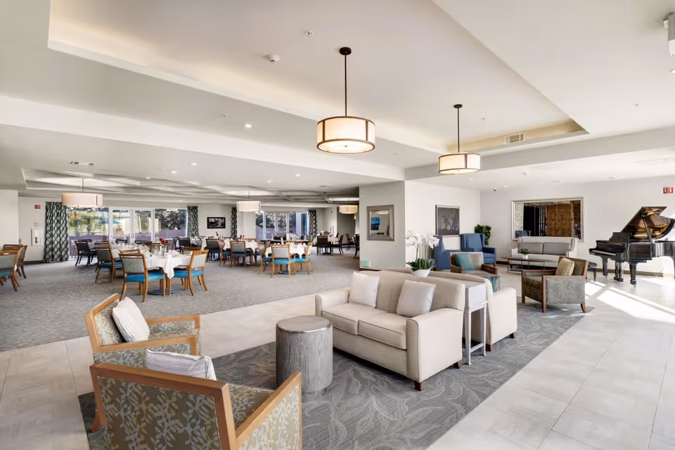 A spacious senior living facility common area featuring a lounge with beige sofas and armchairs, a dining area with multiple tables and chairs, large windows with curtains, and a grand piano near the back wall. The room is well-lit with ceiling lights and natural light from the windows.