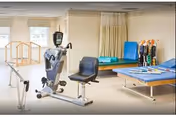 A physical therapy room with exercise equipment including a recumbent bike, parallel bars for walking support, and padded therapy tables with cushions and therapy tools arranged on them. The room has large windows allowing natural light.