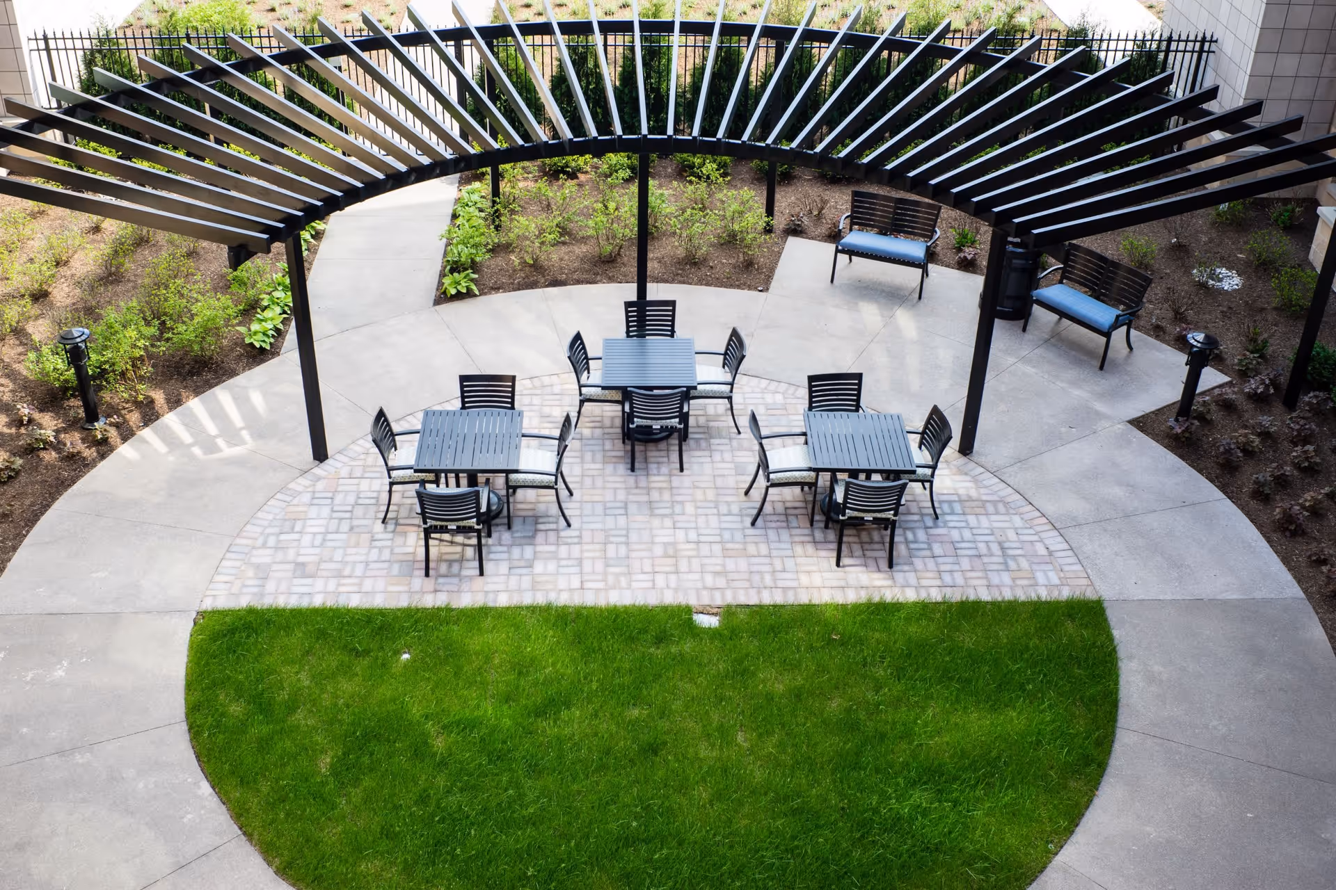 Outdoor seating area with three square tables each surrounded by four black chairs with cushions, under a black pergola structure. There are two benches with blue cushions on a concrete patio surrounded by landscaped garden beds and a green lawn in the foreground.