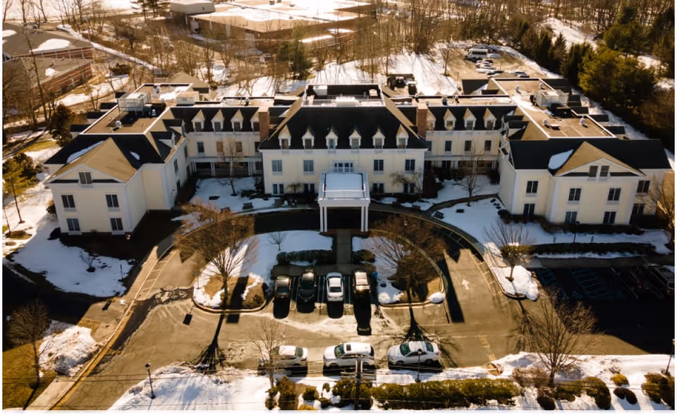 Aerial view of a large, multi-wing building with a circular driveway and parking area in front, surrounded by snow-covered ground and trees in a suburban setting.