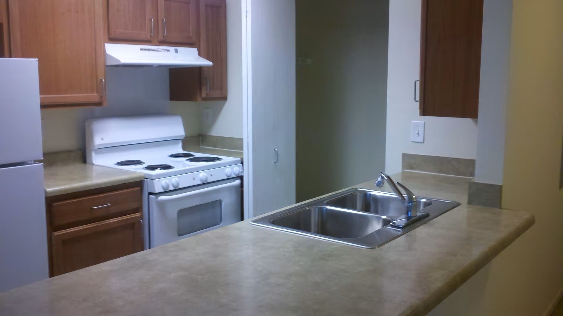 Interior view of a kitchen featuring a white refrigerator, a white electric stove with an oven and range hood, wooden cabinets, and a countertop with a double stainless steel sink and faucet.
