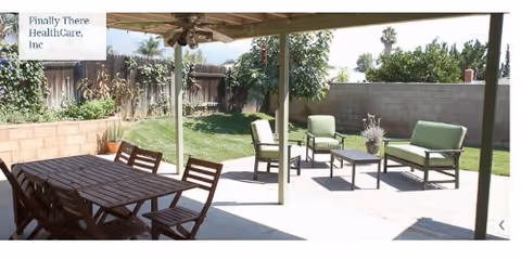 Outdoor patio area with a wooden dining table and chairs under a covered section, and green cushioned seating with a small table on a concrete patio. The area is surrounded by a garden with grass, plants, and a wooden fence.
