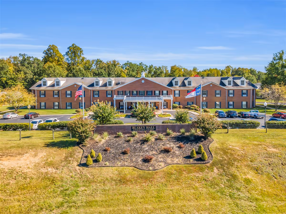 Front exterior view of Blakey Hall Retirement Community, a large brick building with multiple windows and dormers. There are two flagpoles with flags in front of the building, surrounded by landscaped bushes and a parking lot with several cars. Trees and a clear blue sky are visible in the background.