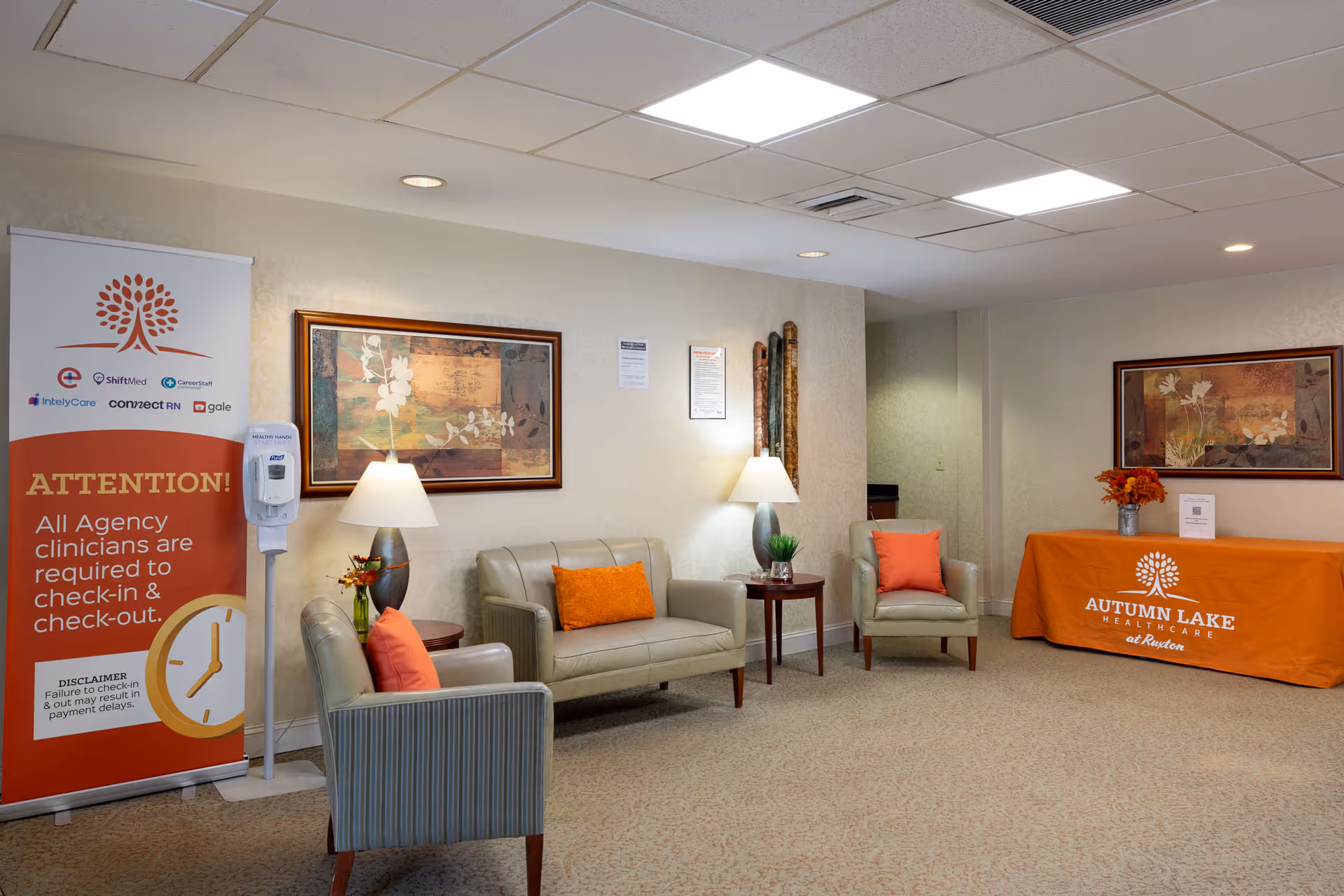 A waiting area in a healthcare facility with beige walls and carpet. The room features a beige loveseat and two armchairs with orange and coral throw pillows. There are two table lamps on small wooden side tables, decorative wall art, and a hand sanitizer dispenser next to a sign instructing agency clinicians to check in and out. A table covered with an orange cloth displays the Autumn Lake Healthcare at Ruxton logo and a vase with orange flowers.