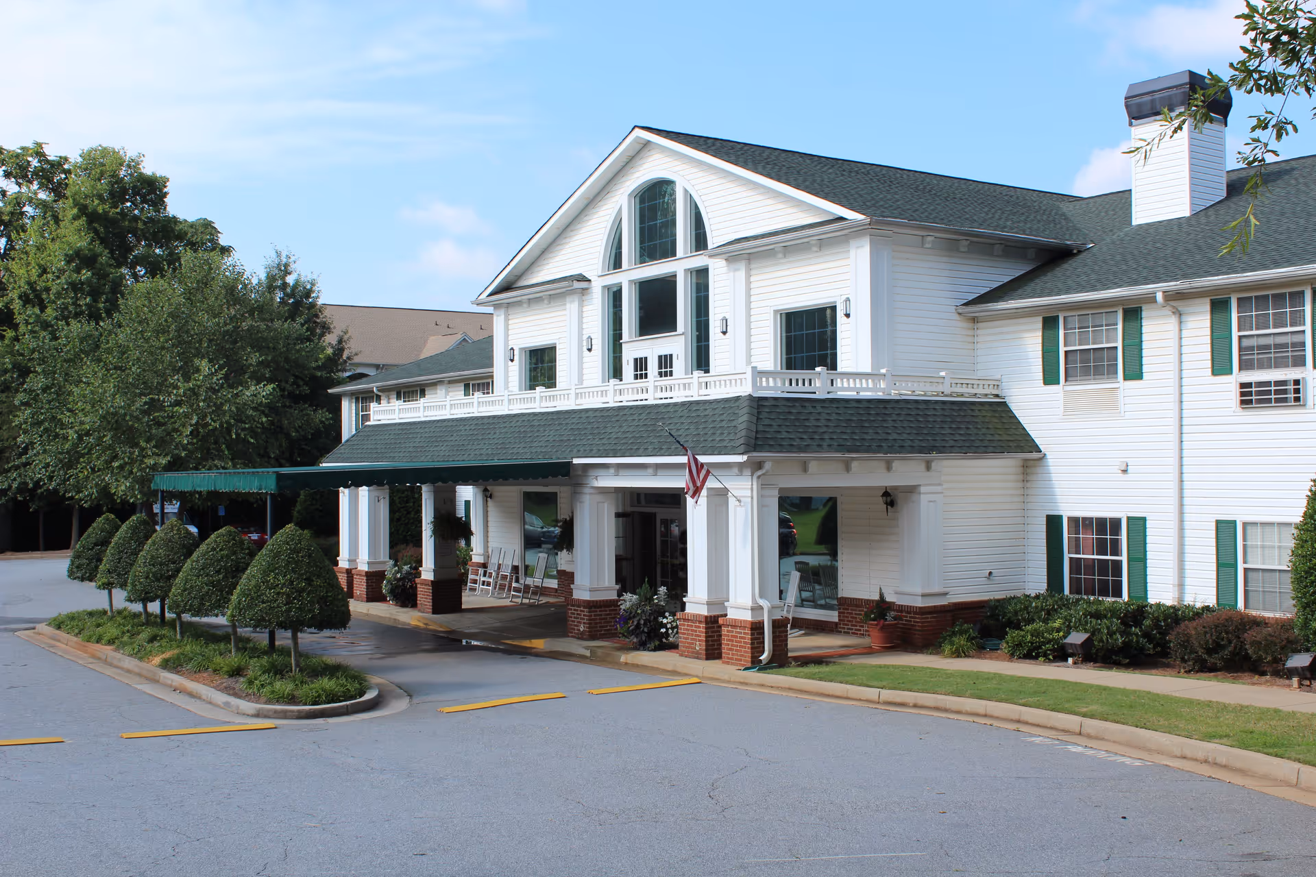 Front exterior view of a two-story white building with green shutters and a green roof. The entrance has a covered driveway with white columns and brick bases, an American flag, and neatly trimmed bushes along the driveway. The sky is clear with a few clouds.