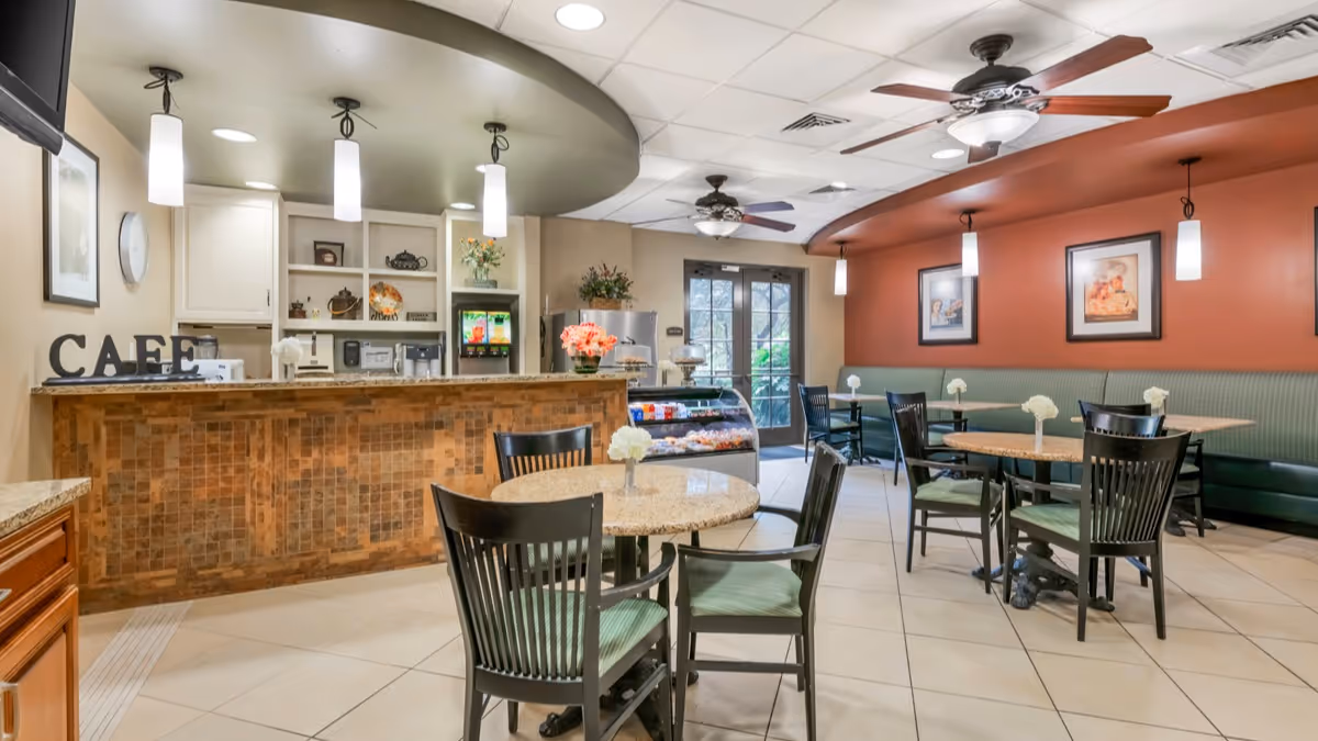 A cozy cafe area inside a senior living facility with a tiled counter, three hanging pendant lights, and a sign that reads 'CAFE'. There are several round tables with green cushioned chairs and small white flower centerpieces. The walls are painted in warm tones with framed artwork, and ceiling fans are visible. A glass door leads outside, allowing natural light to enter the space.
