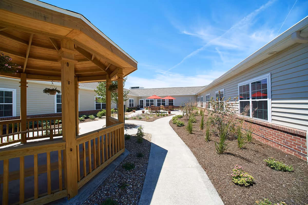 Outdoor courtyard area at Danbury Millersburg featuring a wooden gazebo on the left, a concrete walkway, landscaped garden beds with small plants, and a seating area with tables and red umbrellas in the background under a clear blue sky.