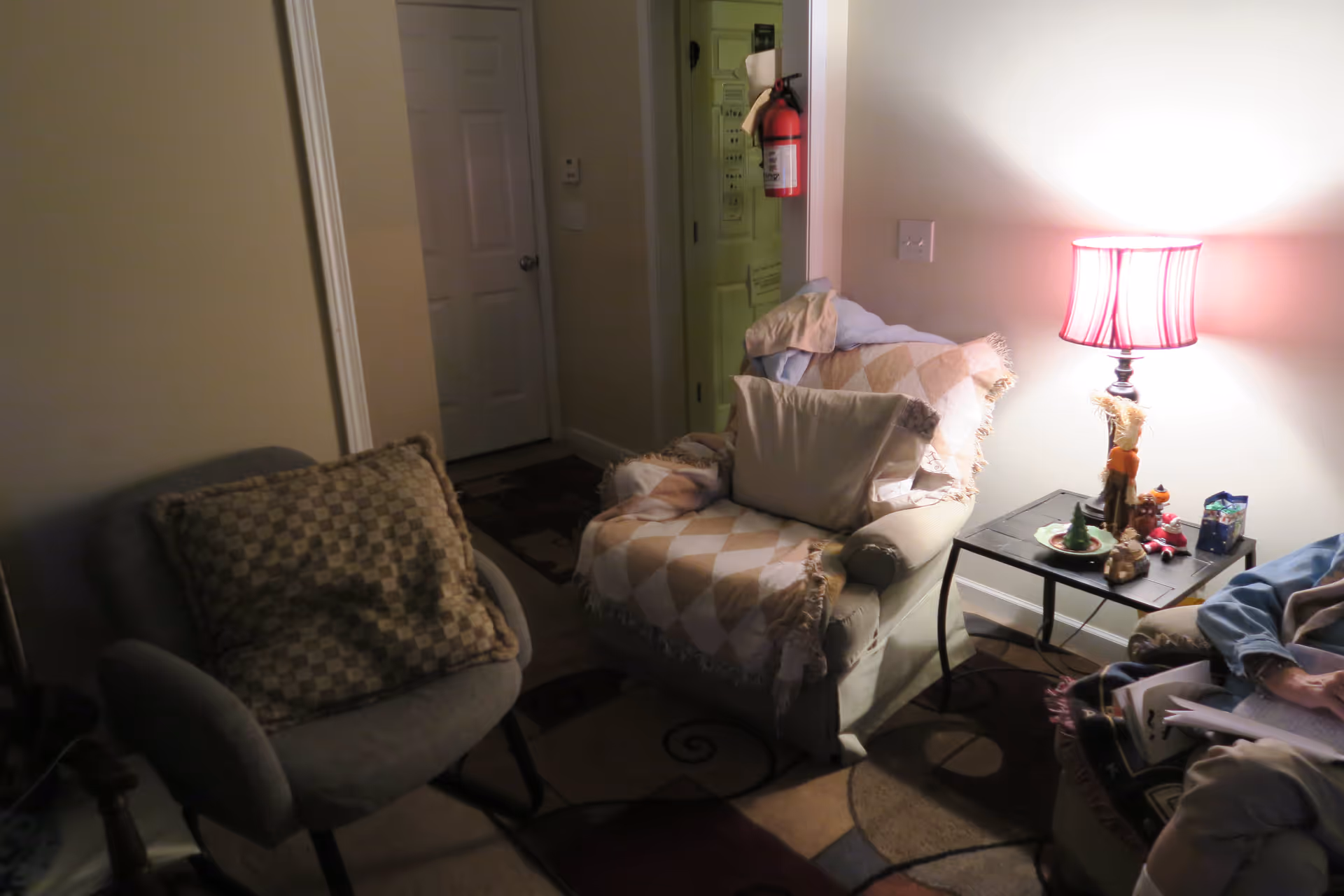 Cozy living room with two upholstered chairs, a side table holding a lamp and small decorations, and a doorway in the background.