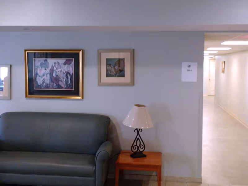 A small seating area with a dark green leather couch and a wooden side table with a decorative lamp on it. Above the couch are three framed pictures hanging on a light gray wall. To the right, there is a hallway with white walls and tiled floor leading to other rooms.