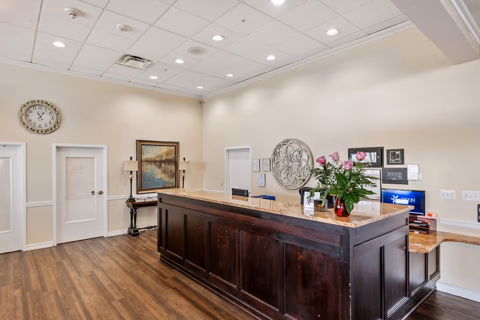 Lobby reception area with a dark wood front desk, a vase of roses, wall clock and decorative artwork.