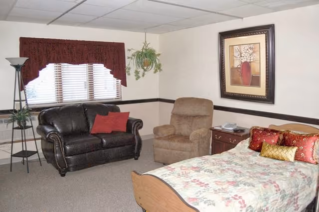 A senior living resident room with a bed, loveseat, recliner, side table, and wall art.