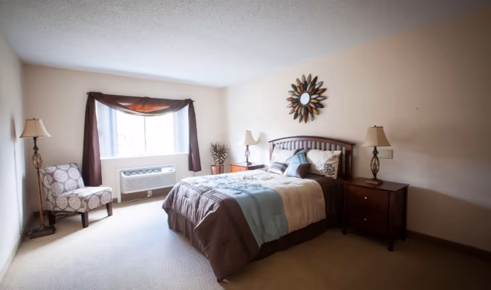 A bedroom with a double bed featuring a brown, beige, and blue comforter, two matching bedside tables with lamps, a decorative mirror above the bed, a patterned armchair near a window with brown curtains, and a wall-mounted air conditioning unit below the window.