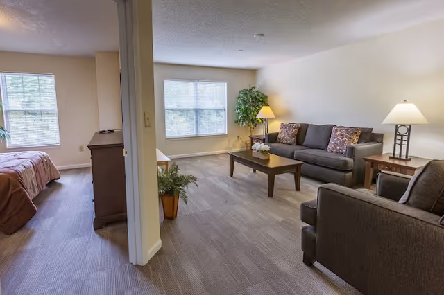 Open-plan living area with sofas, coffee table and lamps, and a bedroom visible through a doorway.