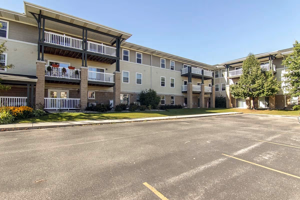 Exterior view of a three-story senior apartment building with balconies, surrounded by a small lawn and trees, and an empty parking lot in the foreground under a clear blue sky.