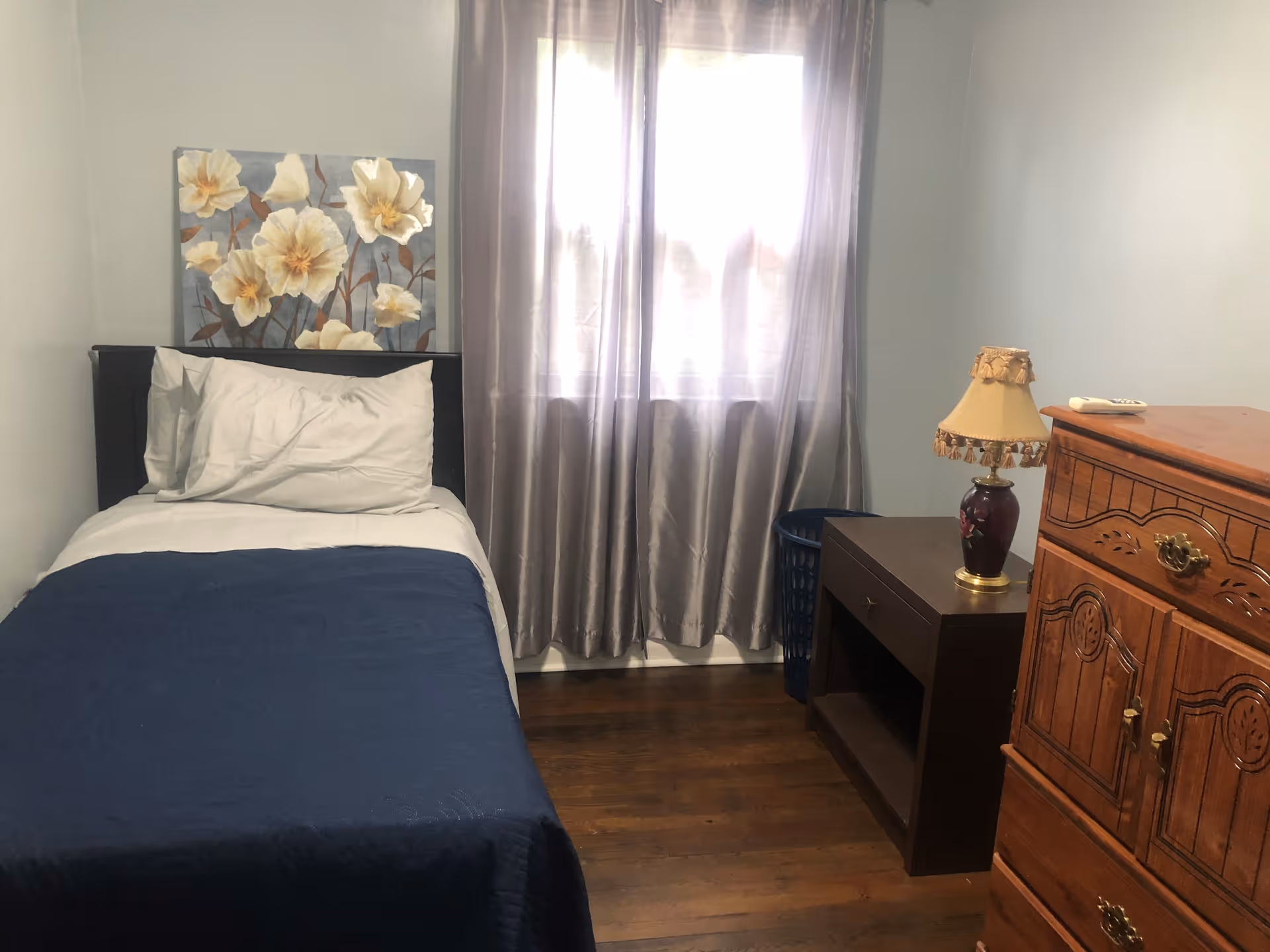 A small bedroom shows a single bed, nightstand, dresser, and a window with sheer curtains.