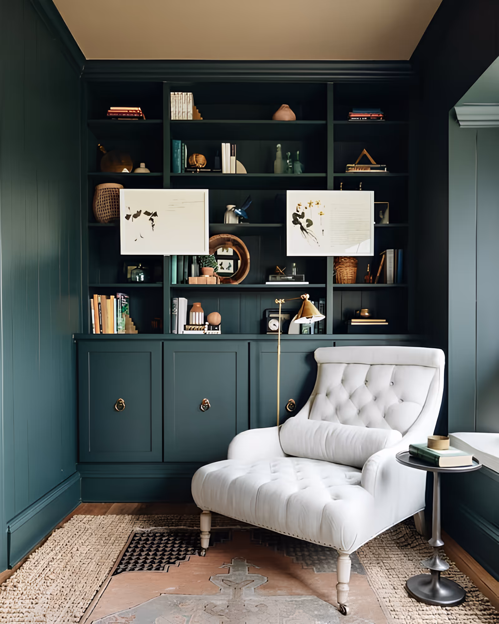 A cozy reading nook featuring a plush white tufted armchair with a cylindrical pillow, positioned next to a small round side table with books and a decorative item. Behind the chair is a dark green built-in bookshelf filled with books, decorative objects, and two abstract art pieces. The floor has a patterned rug, and the walls are painted in a matching dark green color.