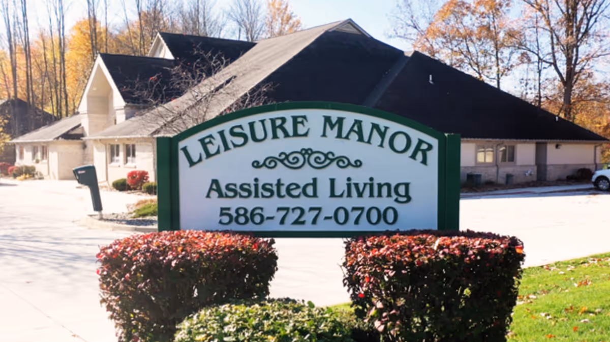Outdoor view of Leisure Manor Assisted Living facility sign with the building and parking area in the background surrounded by trees with autumn foliage.