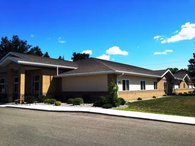 Exterior view of a single-story assisted living and memory care facility building with a brick and siding facade, a sloped roof, and a small landscaped area with bushes and grass under a clear blue sky.