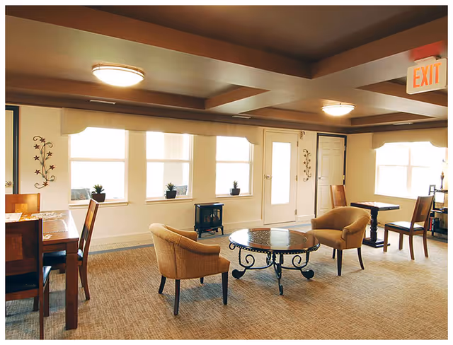 Bright communal lounge with armchairs around a glass-top coffee table, dining table and windows.