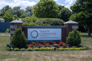 Outdoor view of a brick and stone sign for Trinity Community at Fairborn, surrounded by green shrubs and red flowers, with trees and a blue sky with clouds in the background.