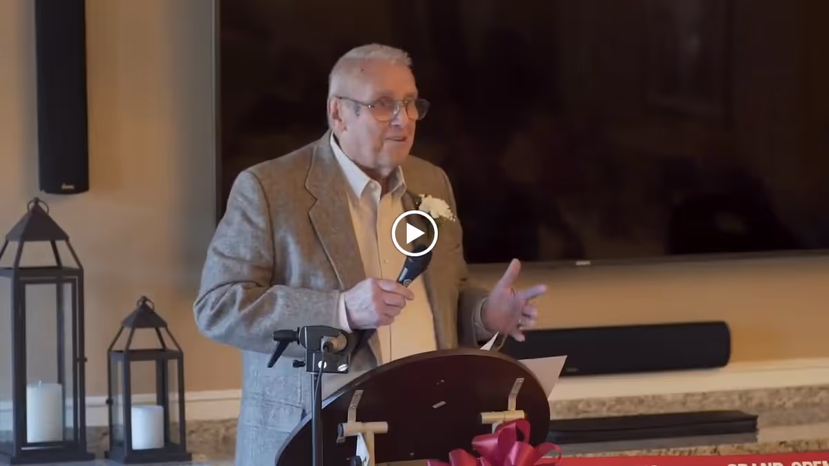An elderly man wearing glasses and a gray blazer is speaking into a microphone while standing behind a podium in a room with a large flat-screen TV mounted on the wall and two decorative lanterns with candles on a countertop.