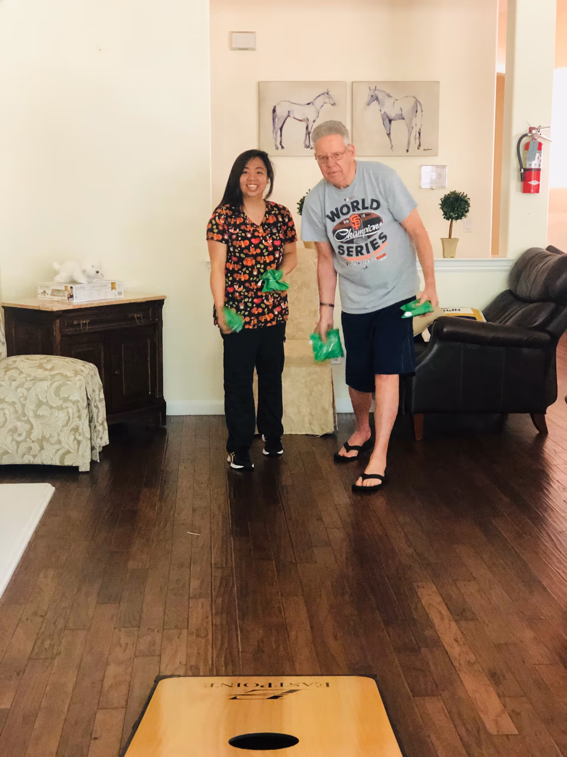 An elderly man and a woman standing indoors on a wooden floor playing a game with green bean bags. The man is wearing a gray World Series t-shirt, black shorts, and flip-flops, while the woman is wearing a floral patterned shirt and black pants. Behind them are two framed pictures of horses on the wall, a small potted plant, a fire extinguisher, a dark brown leather chair, and a wooden cabinet with a white decorative item on top.