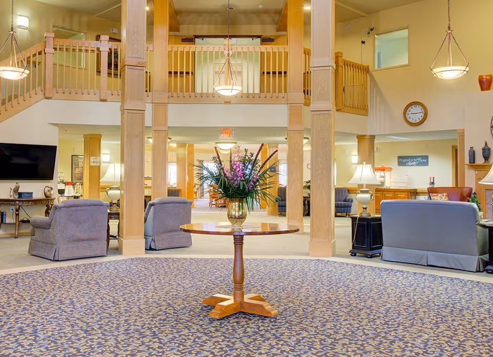 Spacious senior living lobby with a round table and floral arrangement in the center, seating areas and a wooden balcony above.