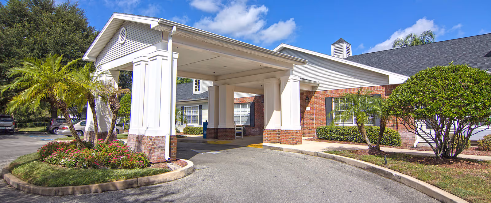 Front exterior view of a senior living facility with a covered entrance supported by white columns, surrounded by landscaped greenery including palm trees and bushes under a blue sky with some clouds.
