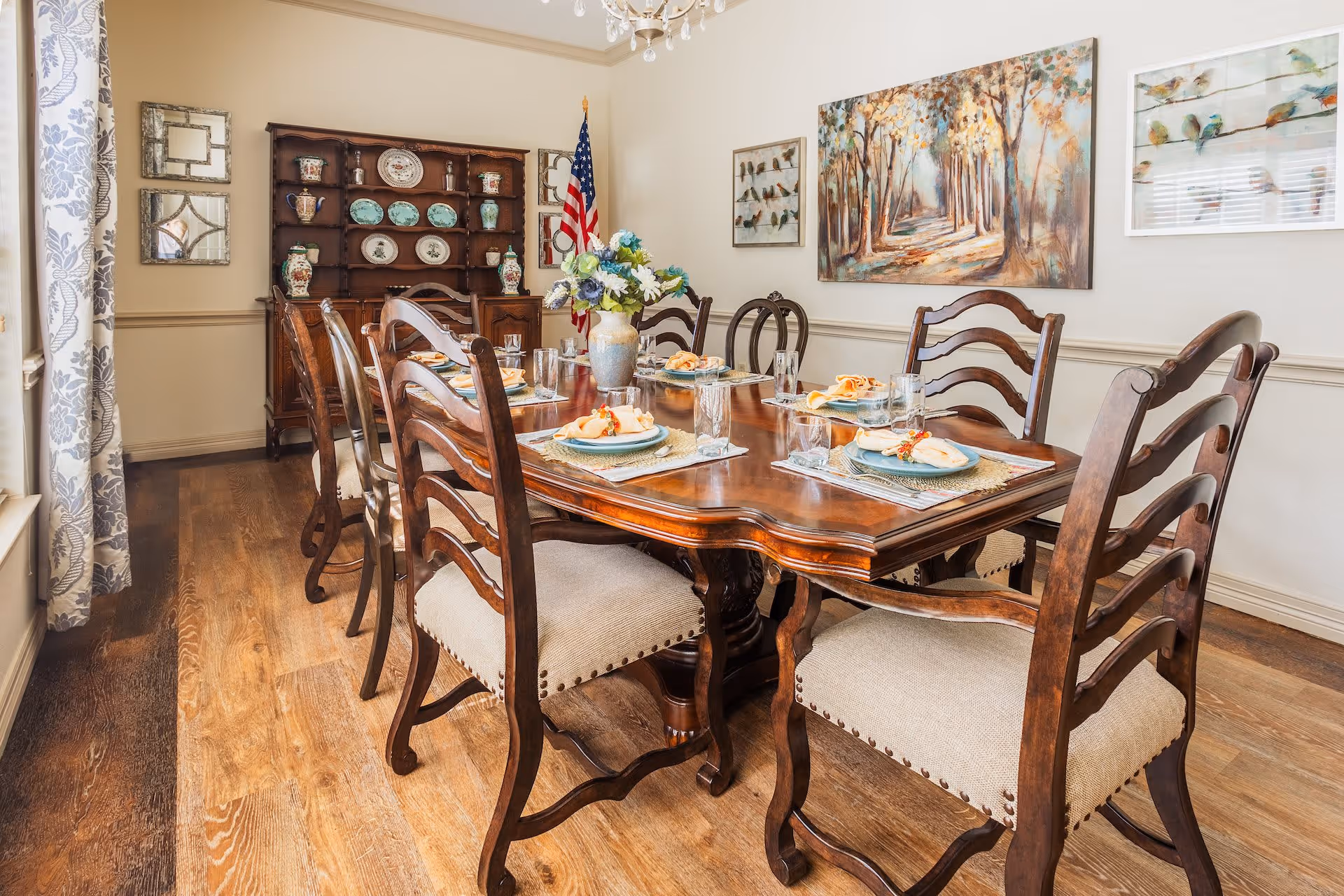 A dining room with a wooden dining table set for six people, featuring plates, glasses, and napkins. The room has wooden flooring, a wooden hutch displaying decorative plates and vases, an American flag, and artwork on the walls including a painting of a tree-lined path and framed bird illustrations. Light-colored walls and patterned curtains frame a window on the left side.