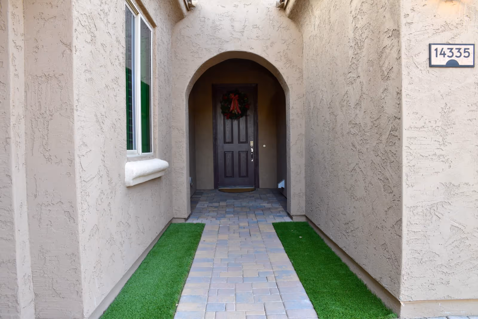 Arched front entryway with a dark wooden door and wreath at house number 14335, flanked by a paver walkway and strips of artificial grass.