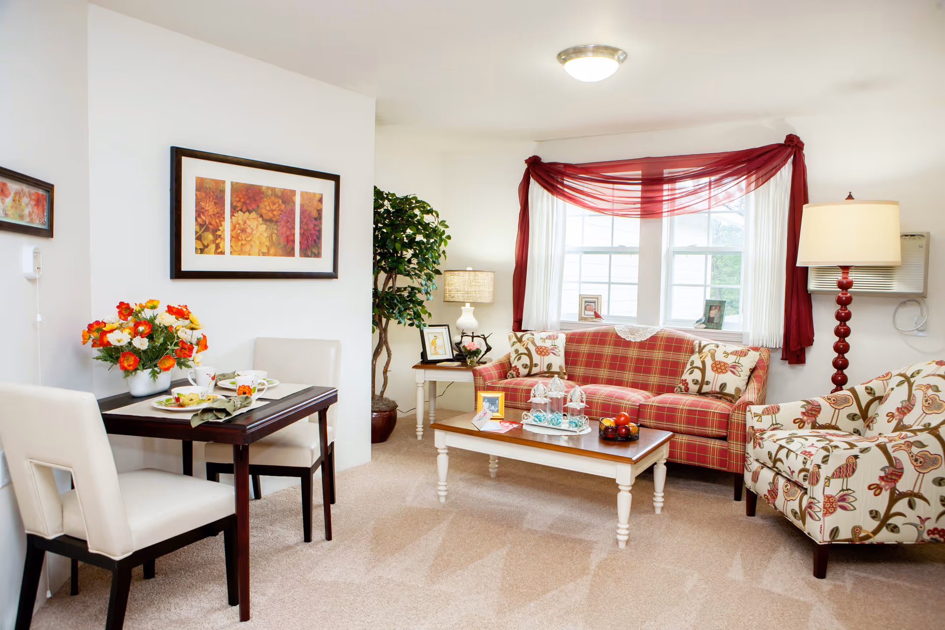 A cozy living room and dining area with a red plaid sofa, floral patterned armchair, white coffee table with decorative items, and a small dark wood dining table with two white chairs. The room has beige carpet, a large window with red sheer curtains, a floor lamp, a table lamp, a potted plant, and framed floral artwork on the wall.