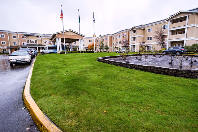 Exterior view of a multi-story senior living facility with beige walls and white trim. The building has balconies and a covered entrance with three flagpoles displaying flags. There is a green lawn and a parking area with several cars parked along the driveway on a rainy day.