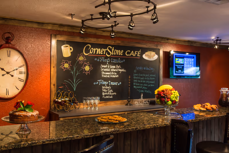 Interior view of CornerStone Café with a granite countertop bar displaying a cake, cookies, and a fruit bowl. Behind the counter is a chalkboard menu listing today's lunch and dinner options, a large wall clock, and a mounted TV screen on a red textured wall.