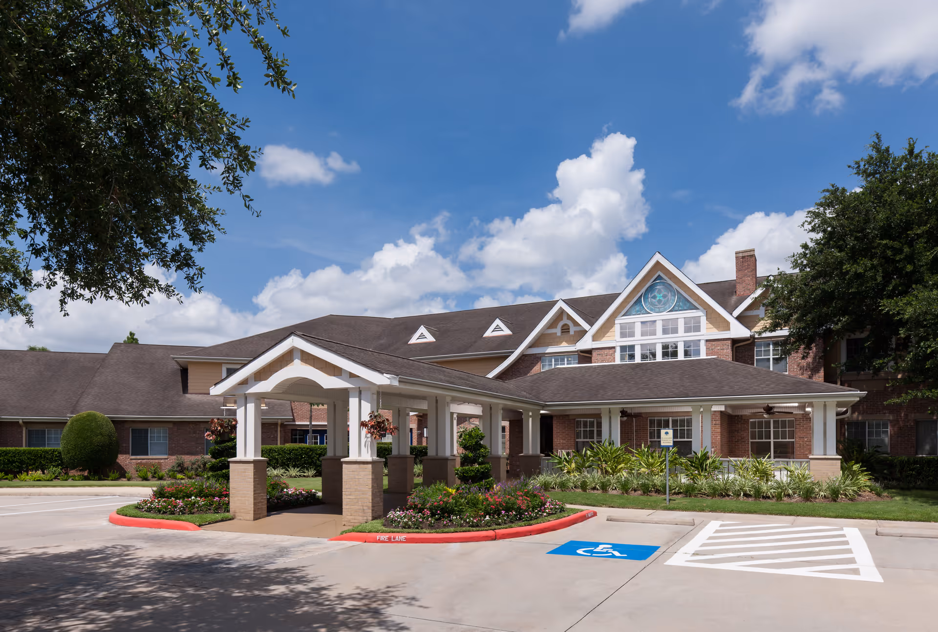 Exterior view of Atria Sugar Land senior living facility showing a large brick building with a covered entrance, landscaped garden beds, and a handicapped parking space under a partly cloudy blue sky.