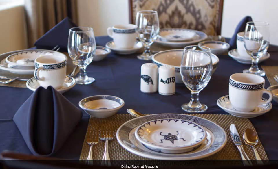A formal dining table set with patterned china, glassware, cups, folded navy napkins and salt and pepper shakers on a dark tablecloth.