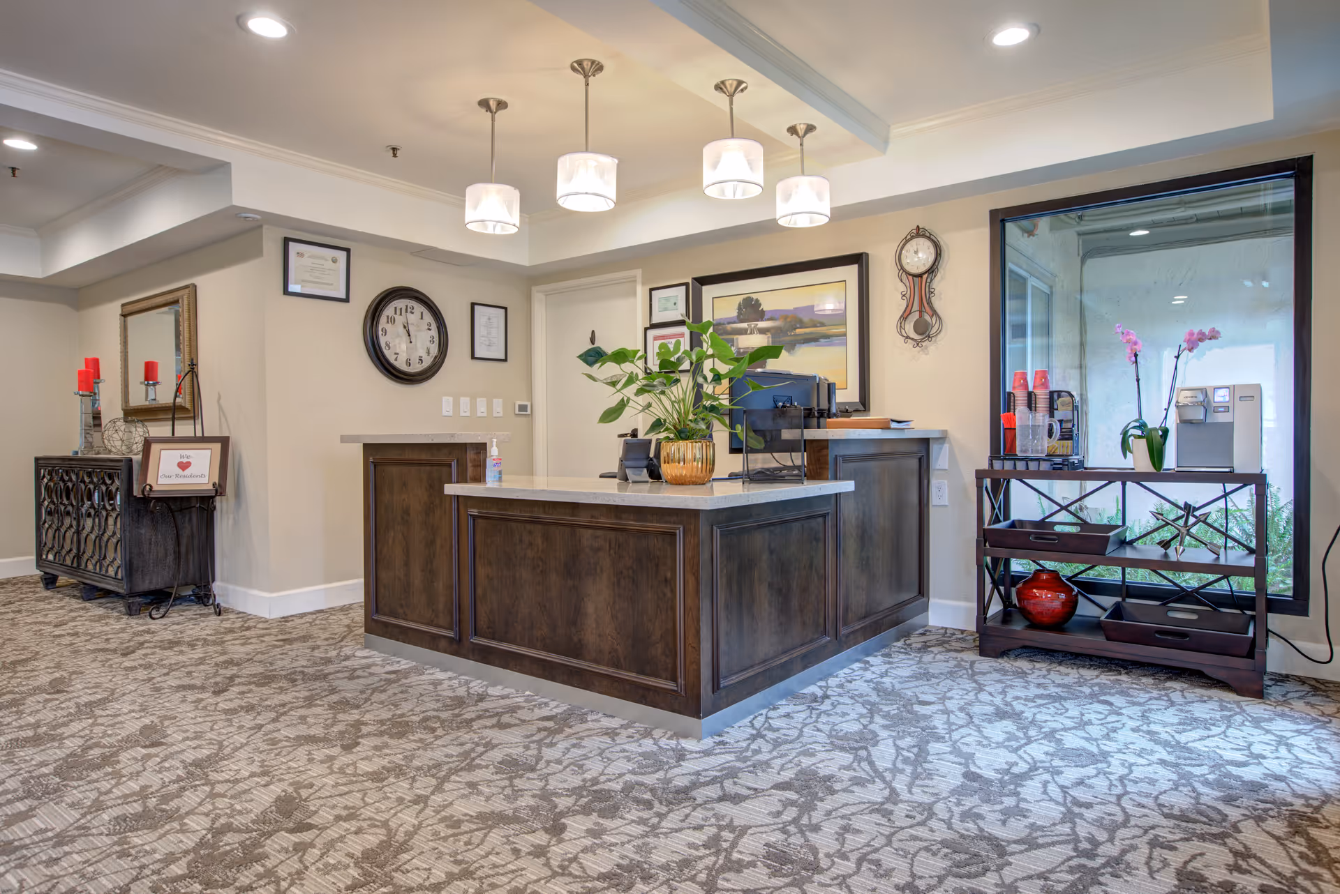 Reception area of BridgePoint at Los Altos featuring a wooden front desk with a plant on top, a wall clock, framed pictures, and a side table with a coffee machine and pink orchids near a large window.