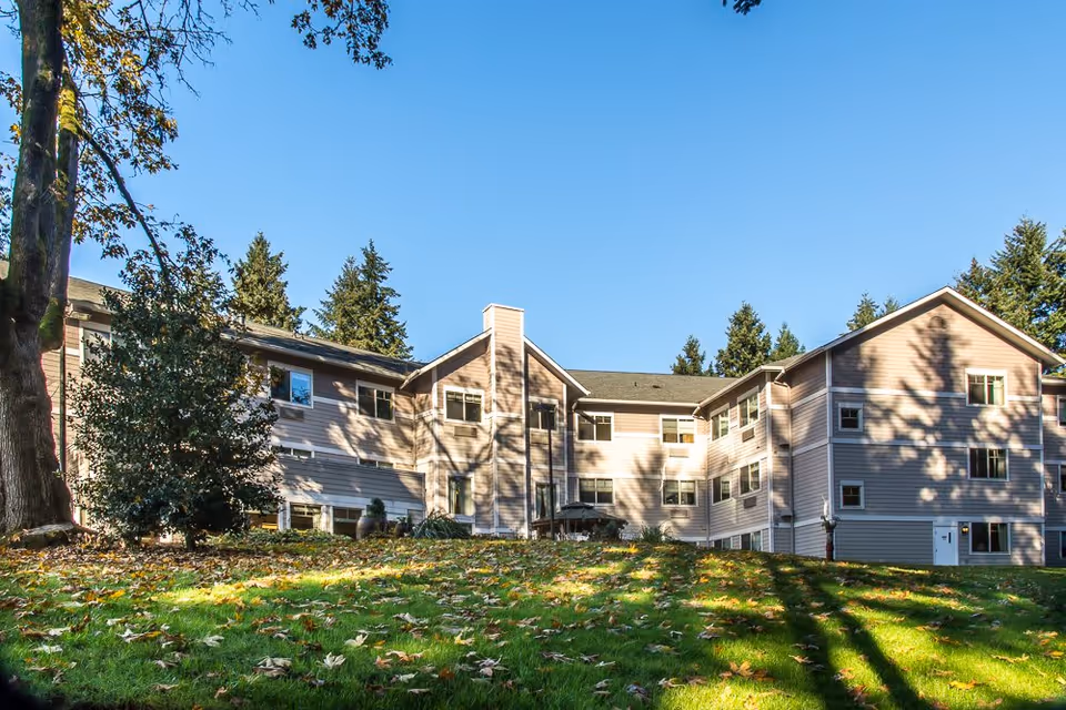 Exterior view of a multi-story assisted living facility building with beige siding, surrounded by trees and a grassy area with fallen leaves under a clear blue sky.