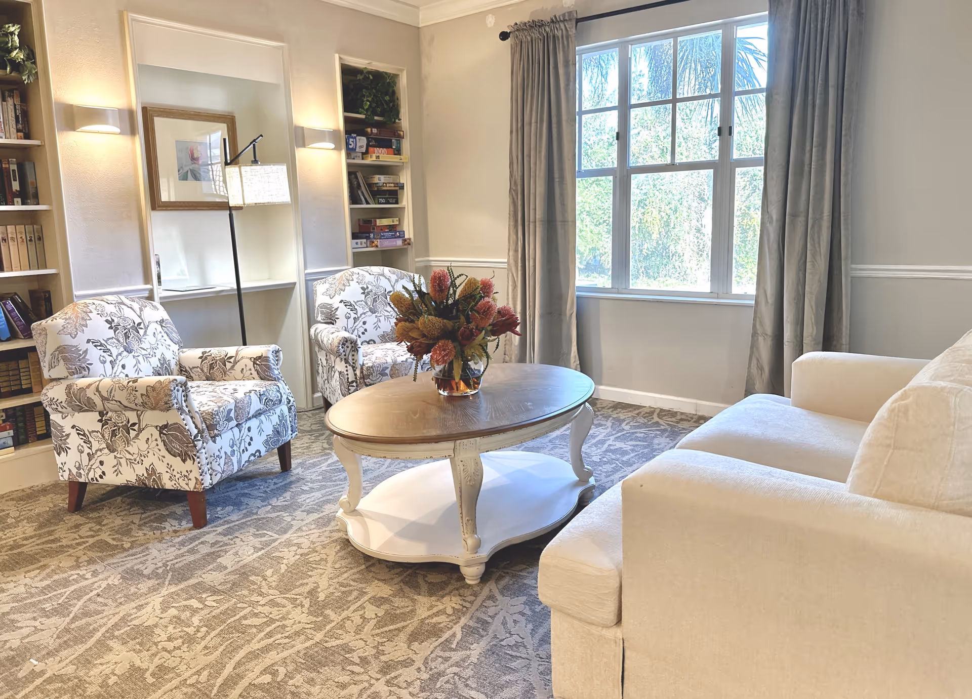 A cozy living room area with a beige sofa, two floral patterned armchairs, and a round wooden coffee table with a floral arrangement. The room has a large window with curtains letting in natural light, built-in bookshelves filled with books and board games, and soft wall lighting.